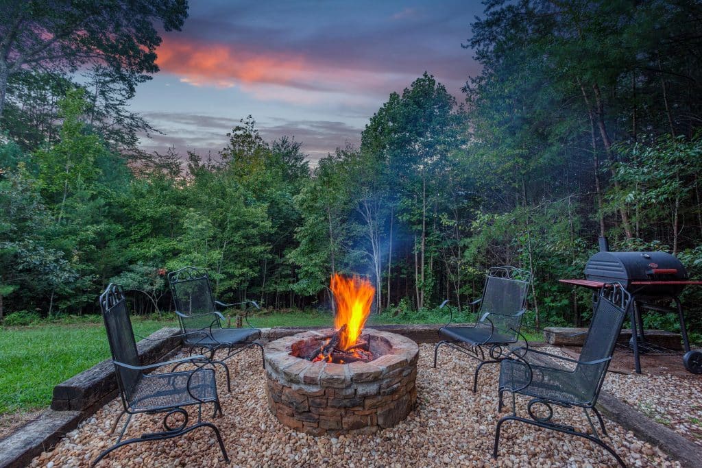 Fire pit with chairs at sunset in forested backyard.