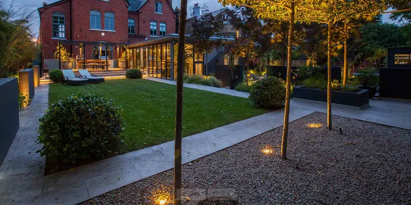 Flush-mounted well light installed in garden gravel to provide architectural grazing on trees without visible hardware.